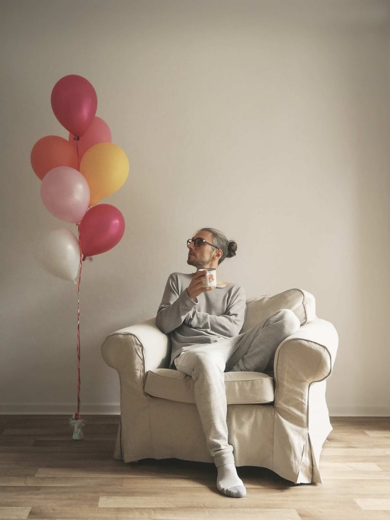 Papajahre auf einem Sessel, daneben schweben bunte Luftballons an schnüren. Er betrachtet die Luftballons. In der Hand eine Tasse.